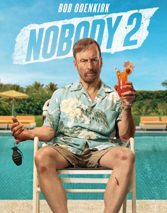 Official movie poster for Nobody 2, featuring Bob Odenkirk as Hutch Mansell, bruised and with cuts on his face, sitting on a poolside chair in a Hawaiian shirt and shorts, holding a grenade in one hand and a tropical drink in the other. The tagline reads "NOBODY RUINS HIS VACATION."