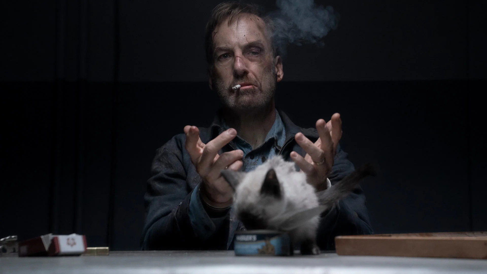 Bob Odenkirk, bruised and smoking, being interrogated while a fluffy white cat on a table from the movie Nobody.