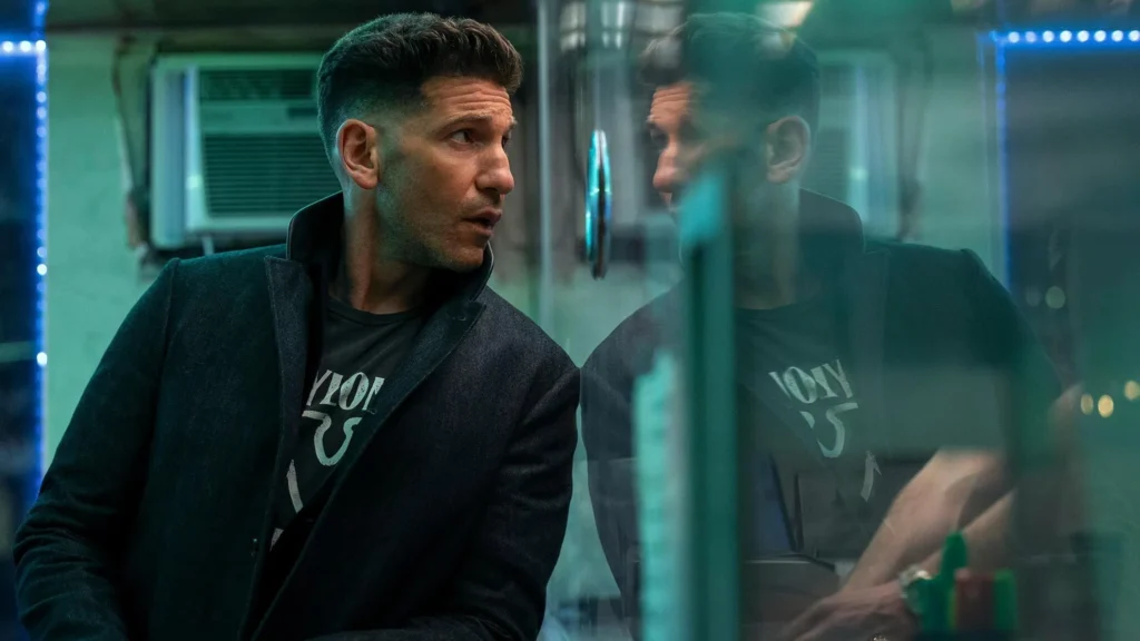 The Accountant 2 Movie Still - Jon Bernthal as Braxton, looking to the side, with his reflection visible in a glass surface in a hotel lobby, bathed in blue and purple neon lights.