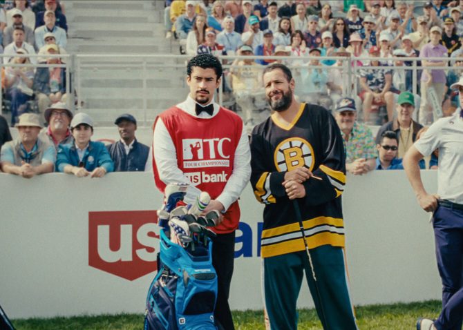 Adam Sandler as Happy Gilmore in his Boston Bruins hockey jersey, holding a golf club, standing next to his caddy on a golf course with spectators in the background.