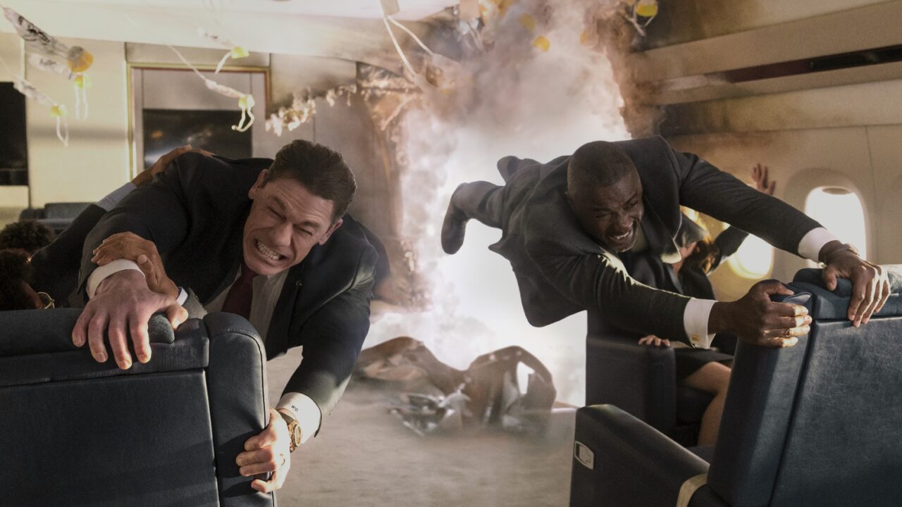 John Cena and Idris Elba, dressed in suits, struggle to hold onto seats in a rapidly disintegrating airplane cabin filled with smoke and debris, their faces strained.