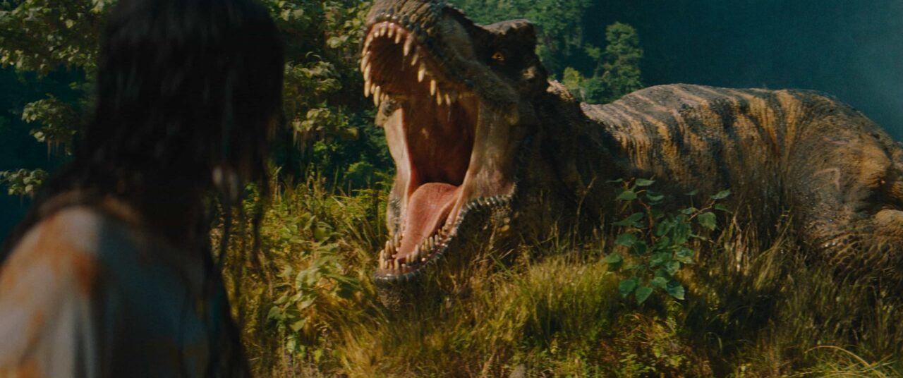 A massive T-Rex roars, jaws wide, at a person with their back to the camera, amidst lush green foliage in a scene from Jurassic World Rebirth.