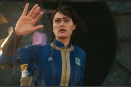 Lucy (Ella Purnell) from Fallout Season 1, wearing a Vault Suit, waving with a look of awe or slight trepidation as she exits a Vault into the wasteland.