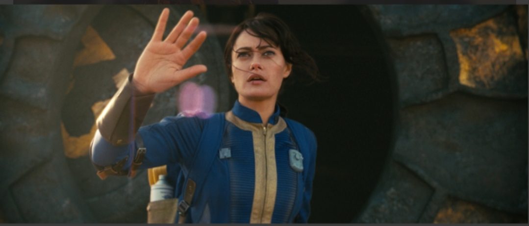 Lucy (Ella Purnell) from Fallout Season 1, wearing a Vault Suit, waving with a look of awe or slight trepidation as she exits a Vault into the wasteland.