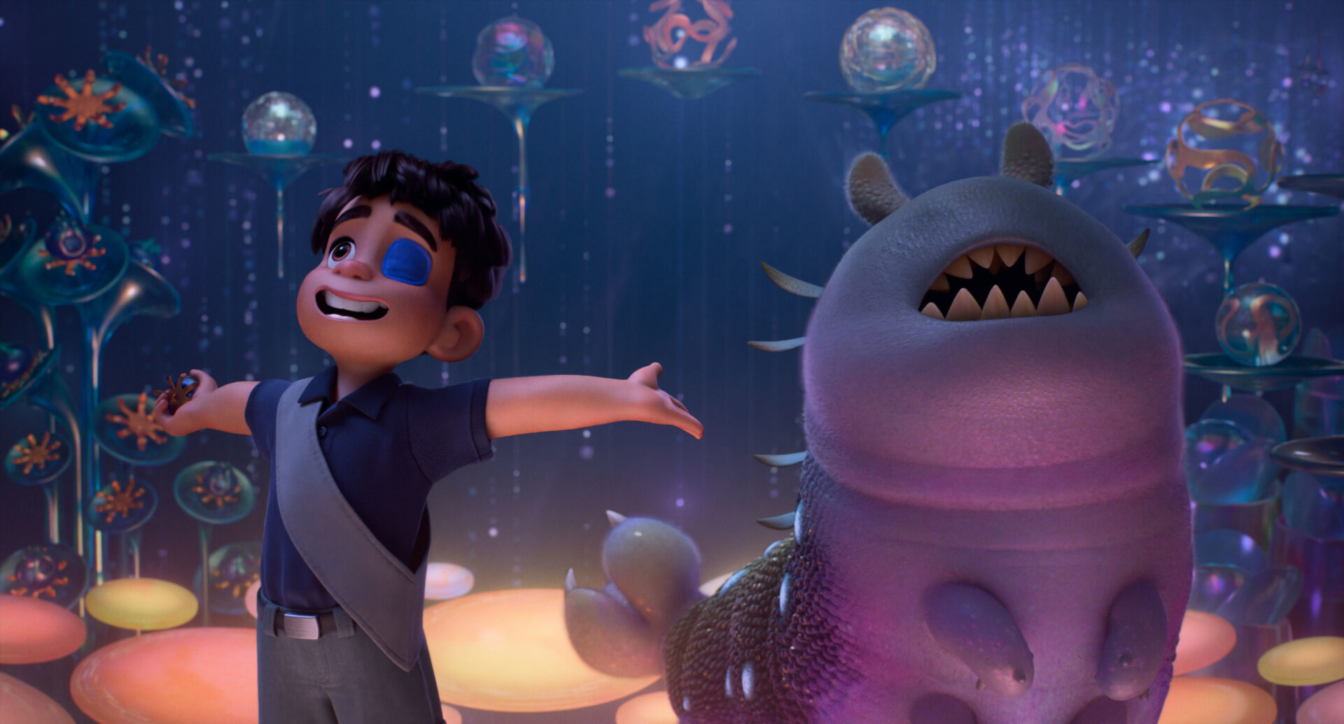 Elio, a young boy with a blue eye patch, stands with arms outstretched next to a large, purple, alien creature with a wide, toothy mouth, in a vibrant, otherworldly setting.