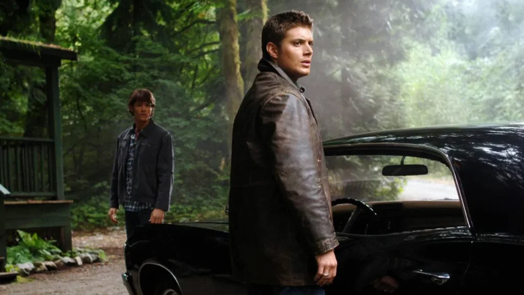 Jensen Ackles (Dean Winchester) wearing a leather jacket, standing next to his 1967 Chevy Impala in a foggy forest with Jared Padalecki (Sam Winchester) in the background.
